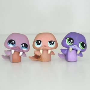 Puede incluir: Tres juguetes de plástico de morsa. Uno es morado, otro es marrón y otro es rosa. Tienen ojos grandes y colmillos.