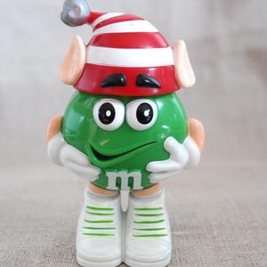 May include: A green M&M character figurine wearing a red and white striped elf hat with a silver pom-pom, pointed ears, white gloves, and white sneakers with green stripes. The character has a smiling face and a white letter m on its torso.