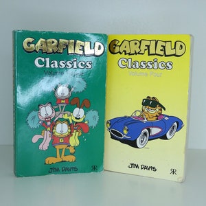 May include: Two paperback books with green and yellow covers. Both books feature the Garfield comic strip character. The book on the left is titled "Garfield Classics Volume One" and the book on the right is titled "Garfield Classics Volume Four".