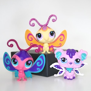 May include: Three colorful, stylized butterfly and animal figurines. The butterfly figurines have large eyes and wings in shades of pink, purple, yellow, and blue. The animal figurine is a light purple color with blue accents.