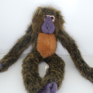 May include: A plush monkey toy with brown fur, a purple face, and orange chest. The monkey is sitting with its arms outstretched.
