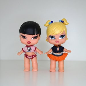 Peut inclure: Deux poupées en plastique aux cheveux noirs et blonds. La poupée aux cheveux noirs porte une chemise rose avec un motif de chat noir et une jupe rouge. La poupée aux cheveux blonds porte une tenue de pom-pom girl bleue et orange avec le numéro 3 sur le devant.