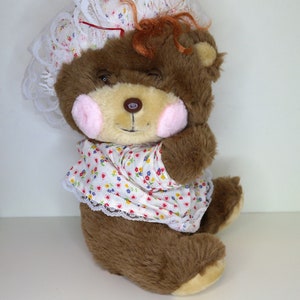 Puede incluir: Un oso de peluche marrón que lleva un gorro de encaje blanco con estampado floral rosa y azul. El oso lleva un vestido blanco con estampado floral rojo, rosa y azul. El oso tiene ojos marrones y una nariz rosa.