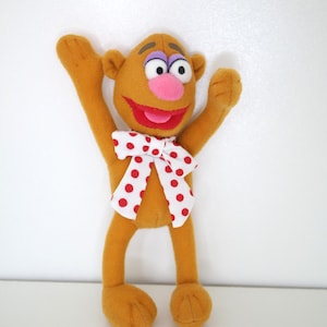 Puede incluir: Un personaje de peluche con cuerpo marrón, brazos levantados y una sonrisa de labios rojos. Tiene una nariz rosa, cejas moradas y una pajarita blanca con lunares rojos. El juguete está sobre una superficie blanca.