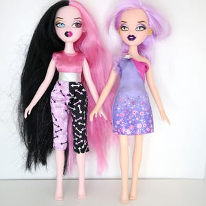 Könnte beinhalten: Zwei Modepuppen mit auffälligem Make-up und Frisuren. Eine Puppe hat schwarzes und rosa Haar und trägt ein rosa Oberteil, einen silbernen Gürtel und eine Hose mit Pfeilmuster. Die andere Puppe hat lila Haare und ein lila Kleid mit floralen Akzenten.