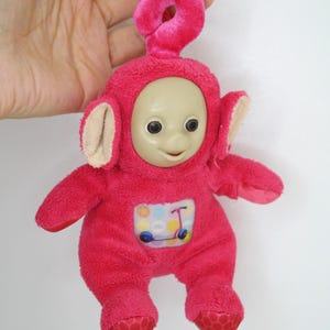 May include: A plush pink Teletubby character with a smiling face, large eyes, and a small scooter graphic on its belly. The toy has a red loop for hanging.