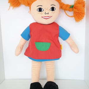 K&ouml;nnte beinhalten: Stoffpuppe mit orangefarbenen Z&ouml;pfen aus Garn, Sommersprossen und einem l&auml;chelnden Gesicht. Die Puppe tr&auml;gt ein rotes Kleid mit gr&uuml;ner Tasche und blauen &Auml;rmeln sowie schwarze Schuhe. Ein klassisches Kinderspielzeug.