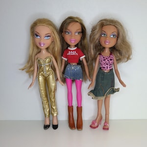 Könnte beinhalten: Drei Bratz-Modepuppen mit blonden, braunen und hellbraunen Haaren. Die Puppen tragen einen goldenen Overall, ein rotes T-Shirt mit dem Text "ROCK ANGELZ" und ein pinkfarbenes Oberteil mit Leopardenmuster. Sie tragen verschiedene Schuhe, darunter schwarze Absätze, braune Stiefel und rosa Sandalen.