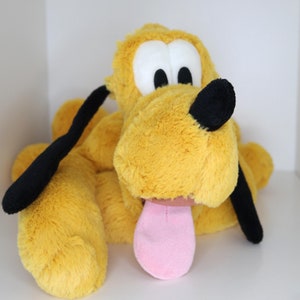 Può includere: Un cane di peluche giallo con orecchie nere e una lingua rosa che sporge. Il cane ha un viso da cartone animato con occhi neri e un naso nero.