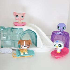 Puede incluir: Un juego de juguetes con figuras de animales. Un cerdo rosa está sentado en un cuenco rosa sobre una estructura blanca cubierta de nieve. Un perro marrón y blanco está sentado en un trineo azul. También se incluye un pingüino en una tabla de surf rosa y un pulpo morado.