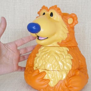 Pode incluir: Um brinquedo de plástico em forma de urso de desenho animado, principalmente laranja e amarelo. O urso tem um nariz azul, um focinho amarelo e um peito amarelo claro. O brinquedo está sobre uma superfície bege.