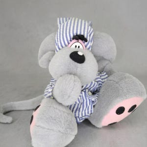 Op de afbeelding: Een pluche grijze cartoonhond met een blauw-wit gestreepte pyjama en een bijpassende nachtmuts. De hond heeft een grote zwarte neus, roze accenten op zijn voeten en een lange staart. Een schattig knuffeldier.