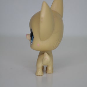 May include: A small, tan-colored plastic toy dog with blue eyes and a pink nose. The dog is standing on a white surface.