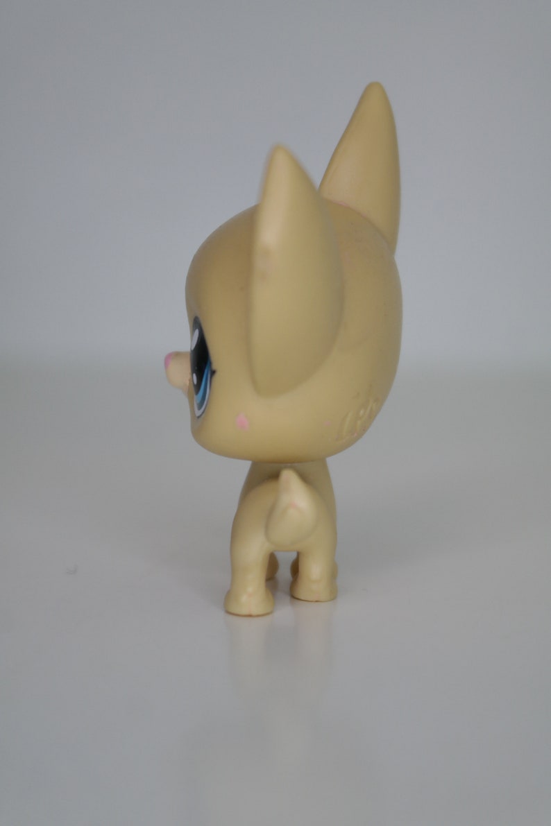May include: A small, tan-colored plastic toy dog with blue eyes and a pink nose. The dog is standing on a white surface.