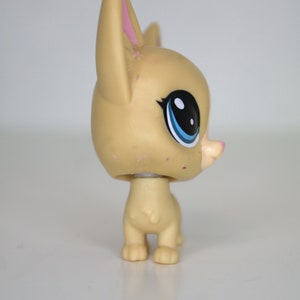 May include: A small, tan-colored plastic toy dog with blue eyes and pink ears. The dog is standing on a white surface.
