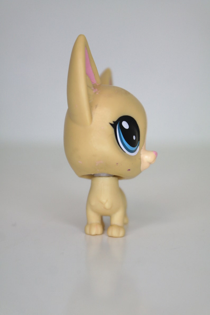 May include: A small, tan-colored plastic toy dog with blue eyes and pink ears. The dog is standing on a white surface.