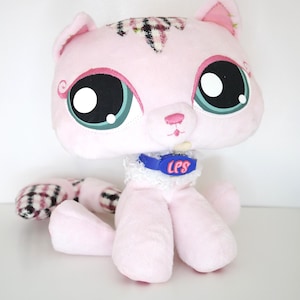 May include: A plush pink cat toy with large black and teal eyes, a blue collar with "LPS" text, and a patterned head and tail. The toy has a white lace collar and is sitting upright.