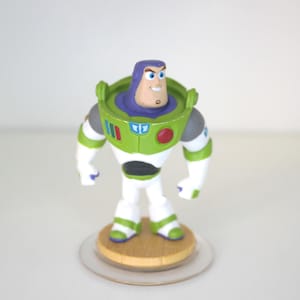 May include: A Buzz Lightyear figure in a white and green spacesuit with purple details. The figure is mounted on a wooden base with a clear plastic disc. The design is based on the character from the Toy Story films.