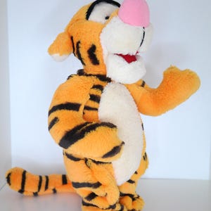 Könnte beinhalten: Ein Plüschtiger mit orangefarbenem Fell und schwarzen Streifen. Der Tiger hat eine rosa Nase, einen weißen Bauch und einen roten Mund. Er steht auf seinen Hinterbeinen und hat die Arme ausgestreckt.
