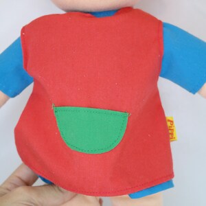 K&ouml;nnte beinhalten: Eine Stoffpuppe mit roten Haaren, Sommersprossen und einem l&auml;chelnden Gesicht. Die Puppe tr&auml;gt ein rotes Kleid mit einer gr&uuml;nen Tasche &uuml;ber einem blauen Hemd. Arme und Beine der Puppe sind sichtbar.