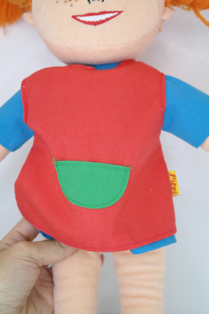 K&ouml;nnte beinhalten: Eine Stoffpuppe mit roten Haaren, Sommersprossen und einem l&auml;chelnden Gesicht. Die Puppe tr&auml;gt ein rotes Kleid mit einer gr&uuml;nen Tasche &uuml;ber einem blauen Hemd. Arme und Beine der Puppe sind sichtbar.