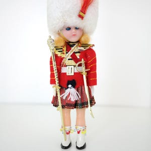 May include: A doll dressed in a red military-style jacket with gold trim, a plaid kilt, and a tall white faux fur hat with a red feather. The doll holds a gold-colored mace and a sword, wearing white boots with colorful accents and a white sporran.