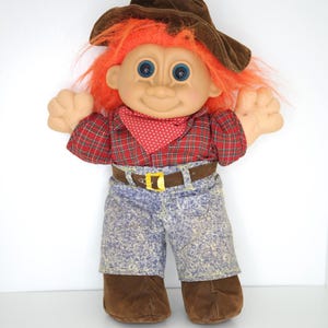 Op de afbeelding: Een trollenpop met feloranje haar en blauwe ogen, gekleed in een bruine hoed, een rood geruit shirt met lange mouwen, een rood met witte stippen sjaaltje, een bruine riem met een gele gesp, een blauwe spijkerbroek met wassing en bruine stoffen schoenen.