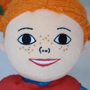 K&ouml;nnte beinhalten: Nahaufnahme einer Pl&uuml;schpuppe mit orangefarbenem Garnhaar, das zu Z&ouml;pfen gestylt ist. Die Puppe hat gestickte Gesichtsz&uuml;ge, darunter gro&szlig;e schwarze Augen, Sommersprossen und einen roten, l&auml;chelnden Mund. Die Puppe tr&auml;gt ein rotes Oberteil mit blauem Kragen.