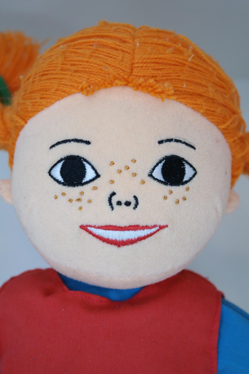 K&ouml;nnte beinhalten: Nahaufnahme einer Pl&uuml;schpuppe mit orangefarbenem Garnhaar, das zu Z&ouml;pfen gestylt ist. Die Puppe hat gestickte Gesichtsz&uuml;ge, darunter gro&szlig;e schwarze Augen, Sommersprossen und einen roten, l&auml;chelnden Mund. Die Puppe tr&auml;gt ein rotes Oberteil mit blauem Kragen.