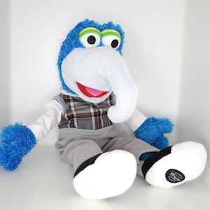 Puede incluir: Un peluche de Gonzo de Los Muppets, con una gran nariz blanca, pelaje azul y ojos grandes verdes y negros. Lleva un chaleco a cuadros, pantalones grises y zapatos blancos con detalles negros.