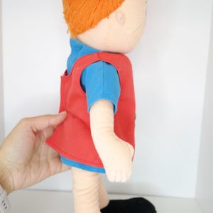 K&ouml;nnte beinhalten: Handgemachte Puppe mit orangefarbenen Garnz&ouml;pfen, die ein blaues T-Shirt und eine rote Weste tr&auml;gt. Die Puppe hat schwarze Schuhe und ist aus weichem Stoff gefertigt. Die Puppe ist ein Kinderspielzeug.