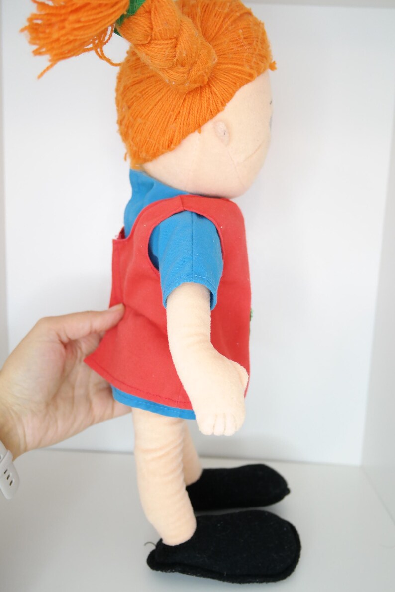 K&ouml;nnte beinhalten: Handgemachte Puppe mit orangefarbenen Garnz&ouml;pfen, die ein blaues T-Shirt und eine rote Weste tr&auml;gt. Die Puppe hat schwarze Schuhe und ist aus weichem Stoff gefertigt. Die Puppe ist ein Kinderspielzeug.
