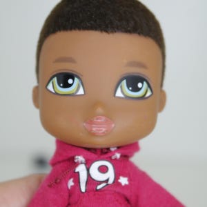 Könnte beinhalten: Eine Puppe mit braunen Haaren und brauner Haut, die ein rosa Kapuzenpullover mit weißen Sternen und der Nummer 19 auf der Vorderseite trägt. Die Puppe hat grüne Augen und trägt eine blaue Jeans.