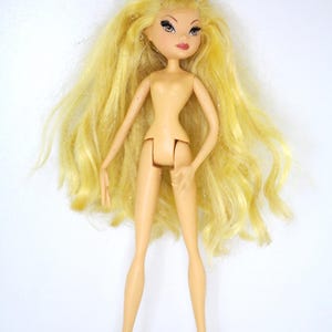 Peut inclure: Une poupée mannequin avec de longs cheveux blonds et une peau bronzée. La poupée a un physique mince, avec des bras et des jambes articulés. Le visage de la poupée présente un maquillage détaillé, notamment des cils foncés et du rouge à lèvres rouge. La poupée est debout sur un fond blanc.