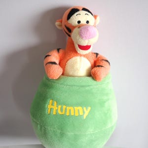 Könnte beinhalten: Ein Plüschtier von Tigger, das aus einem grünen Topf mit dem gelb gestickten Wort "Hunny" herausragt. Das Spielzeug hat orangefarbenes Fell mit schwarzen Streifen, eine rosa Nase und einen roten Mund. Der Topf hat eine weiche, abgerundete Form.