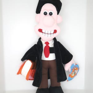 Puede incluir: Un juguete de peluche de un personaje que lleva una toga de graduación negra y un gorro, sosteniendo un libro. El personaje tiene una nariz grande, ojos grandes y una amplia sonrisa. El juguete tiene una corbata roja y pantalones marrones. La etiqueta dice "Wallace & Gromit".