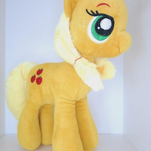 Puede incluir: Un peluche de poni amarillo con crin y cola de color amarillo claro. El poni presenta grandes ojos verdes bordados con pestañas largas y tres iconos de manzanas rojas en el flanco. El juguete tiene una textura suave y se sostiene sobre cuatro patas.