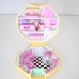 May include: A vintage yellow and pink miniature dollhouse playset, open to reveal furnished rooms. The interior features a bedroom, kitchen, and living area with tiny figures. The floor has a black and white checkered pattern.
