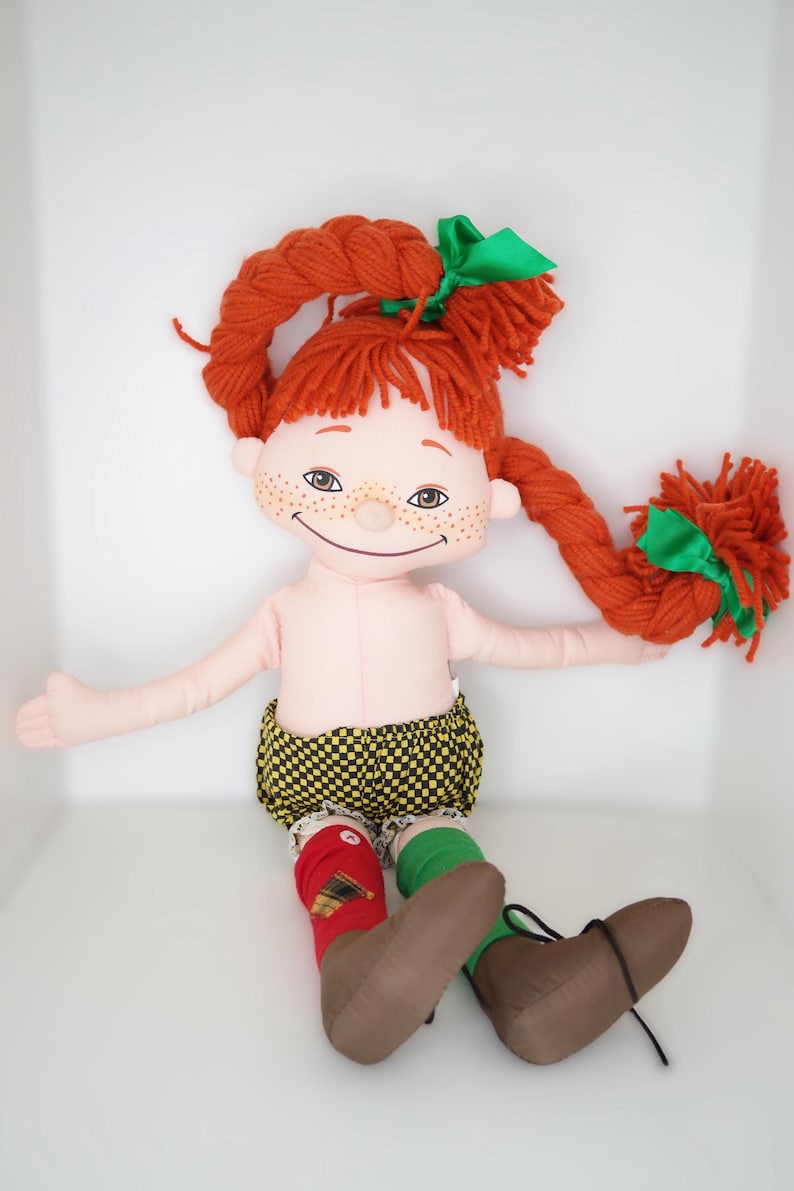 以下が含まれることがあります： 緑のリボンで結ばれた赤い糸の三つ編みを持つ手作りのぼろきれ人形。人形は薄いピンク色の体、そばかすがあり、黄と黒のチェック柄のショーツ、赤と緑の靴下、茶色のブーツを履いています。人形は腕を広げて座っています。