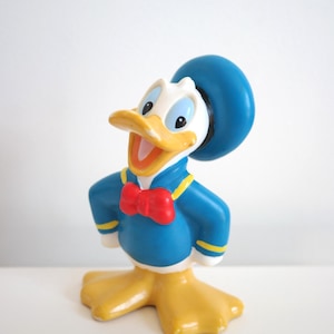 Op de afbeelding: Een geel en blauw plastic beeldje van Donald Duck met een blauwe matrozenpet en een rode strik.