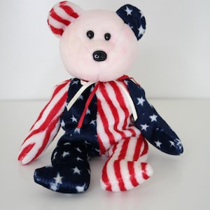 May include: A plush teddy bear with patriotic colors. The bear's body is split with red and white stripes and navy blue with white stars. The ears are red and white striped, and the face is light pink with black eyes and nose.