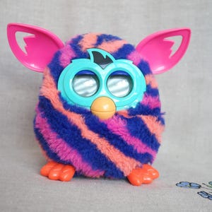 Puede incluir: Un juguete electrónico de peluche con un cuerpo redondo cubierto de pelaje a rayas azules y rosas. Cuenta con grandes orejas de plástico rosa brillante, una placa frontal de plástico azul claro que rodea dos ojos circulares, un pequeño pico naranja y pies naranjas.