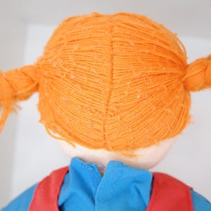 K&ouml;nnte beinhalten: Eine Puppe mit orangefarbenem Garnhaar, das zu zwei Z&ouml;pfen gestylt ist. Die Puppe tr&auml;gt ein blaues Hemd und eine rote Weste. Das Haar der Puppe ist mit gr&uuml;nen B&auml;ndern gebunden.