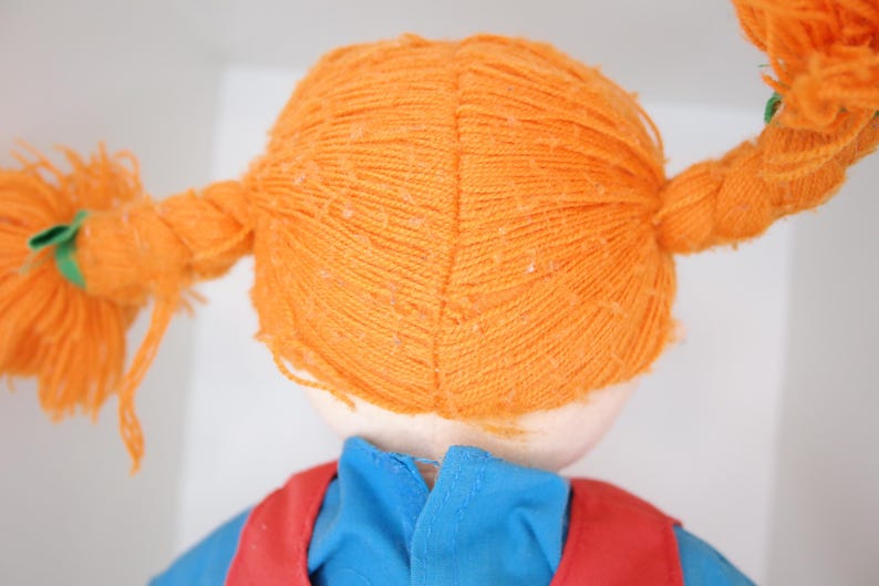 K&ouml;nnte beinhalten: Eine Puppe mit orangefarbenem Garnhaar, das zu zwei Z&ouml;pfen gestylt ist. Die Puppe tr&auml;gt ein blaues Hemd und eine rote Weste. Das Haar der Puppe ist mit gr&uuml;nen B&auml;ndern gebunden.