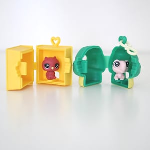 Puede incluir: Dos figuras de animales de juguete en estuches de transporte amarillos y verdes. Un estuche está abierto, revelando una figura de búho rojo. El otro estuche está abierto, revelando una figura de tortuga rosa. Los estuches tienen detalles de corazón y cadena.