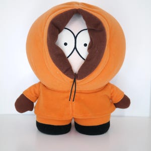 Puede incluir: Muñeco de peluche naranja de un personaje de dibujos animados con ribete de capucha marrón y detalles en negro. El muñeco tiene ojos grandes y blancos y un detalle de cuerda negra. El atuendo incluye una chaqueta naranja y pantalones negros.