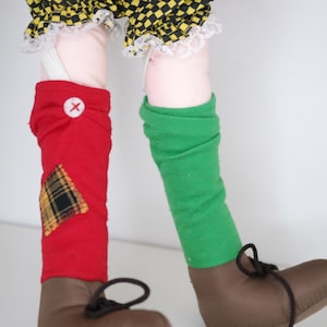 以下が含まれることがあります： 茶色の靴と黒い靴紐、赤と緑のレッグウォーマー、黄と黒のチェック柄のショートパンツを履いた人形の足。赤いレッグウォーマーには、チェック柄のパッチと白いボタンが付いています。