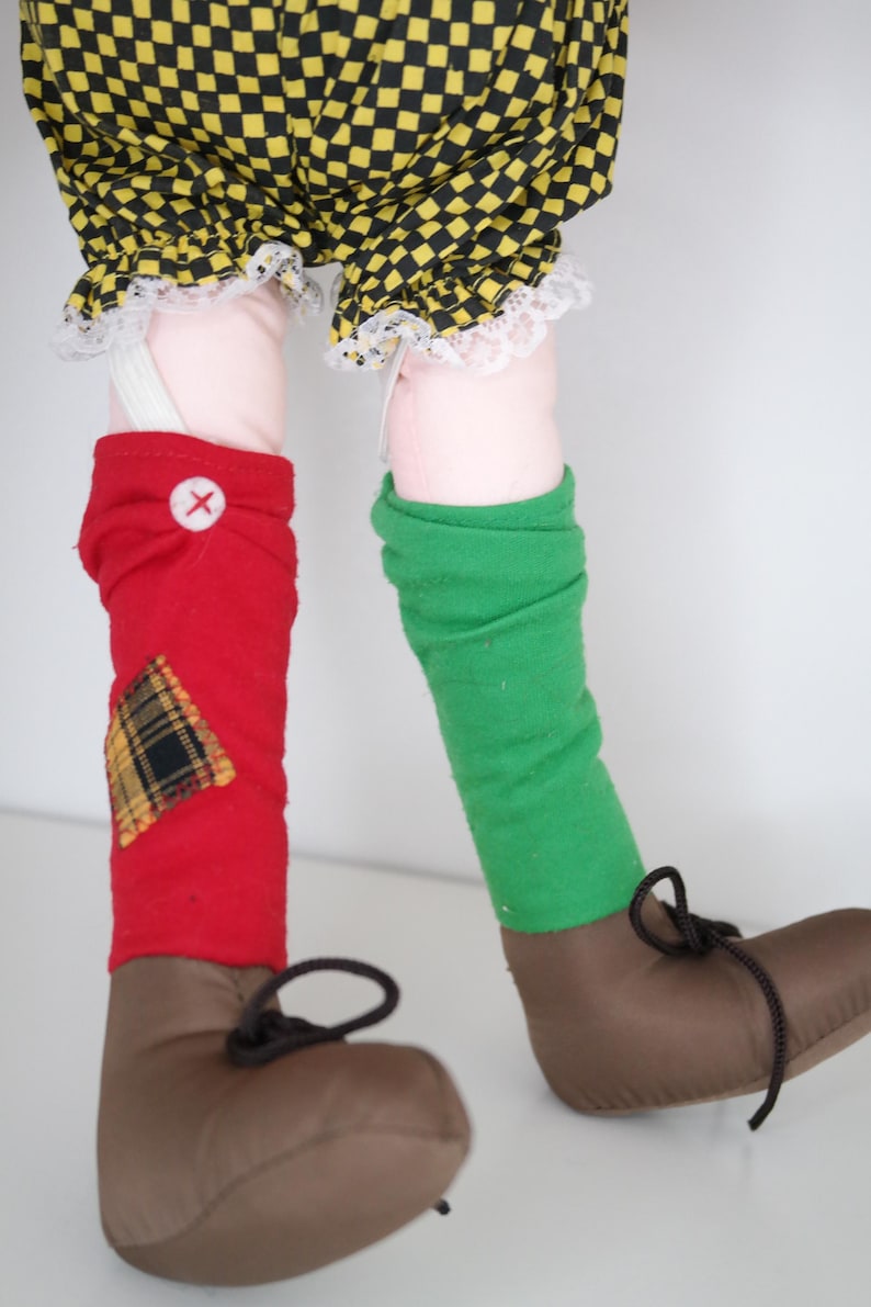 以下が含まれることがあります： 茶色の靴と黒い靴紐、赤と緑のレッグウォーマー、黄と黒のチェック柄のショートパンツを履いた人形の足。赤いレッグウォーマーには、チェック柄のパッチと白いボタンが付いています。