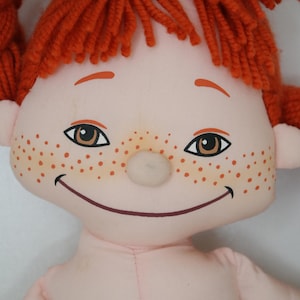 以下が含まれることがあります： 笑顔、そばかす、オレンジ色の糸のおさげ髪が特徴の手作りの人形。人形は茶色の目をし、肌の色はピンク色です。頭の上には緑のリボンが結ばれています。人形の体は薄いピンク色です。