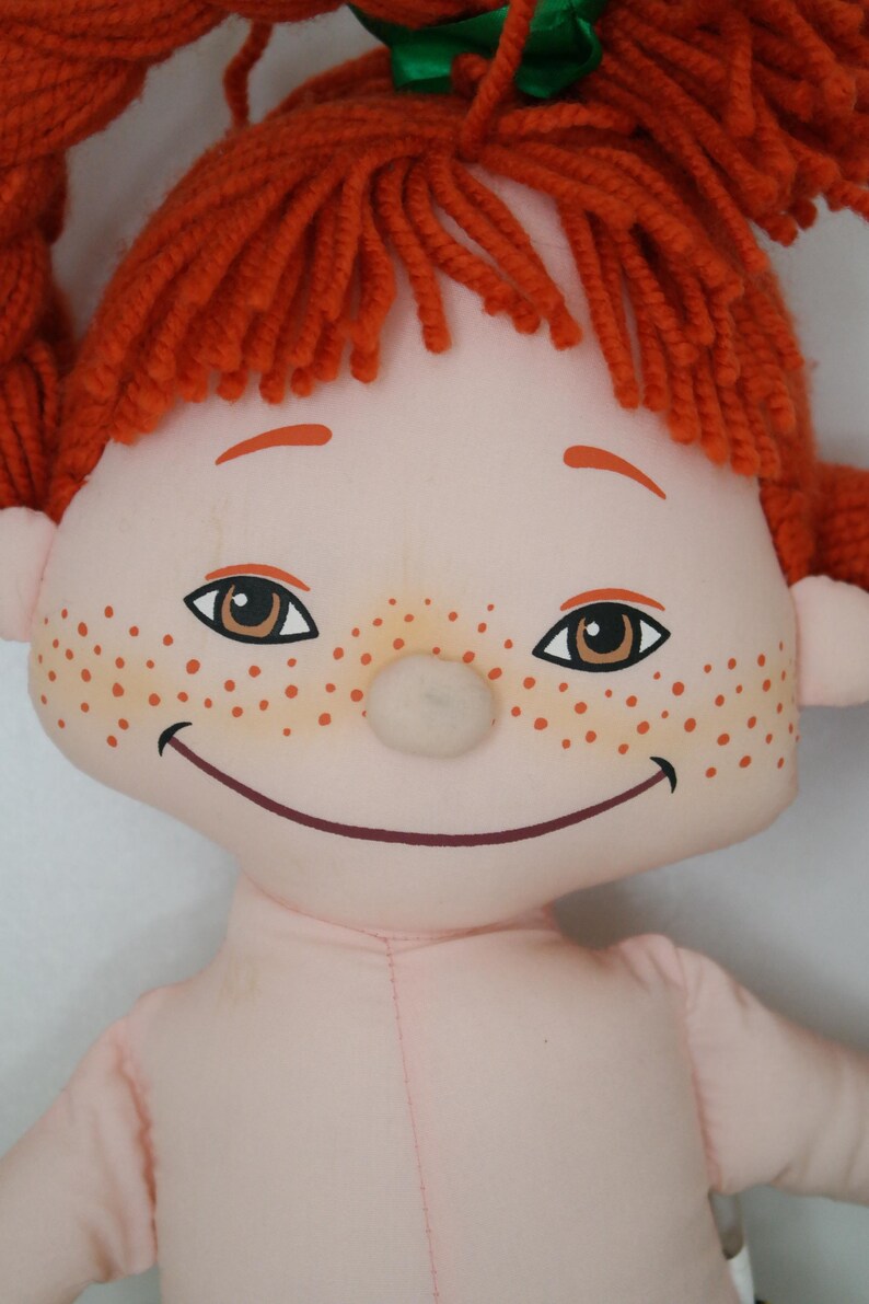 以下が含まれることがあります： 笑顔、そばかす、オレンジ色の糸のおさげ髪が特徴の手作りの人形。人形は茶色の目をし、肌の色はピンク色です。頭の上には緑のリボンが結ばれています。人形の体は薄いピンク色です。